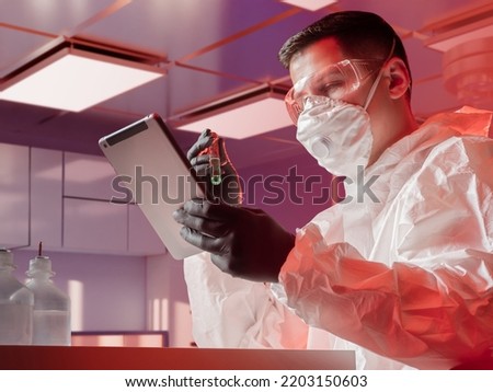 Man virologist. Virologist in protective white suit. Guy with tablet and glass flask. Young man works in laboratory. Virologist studies dangerous pathogen. Researcher at virology laboratory Royalty-Free Stock Photo #2203150603