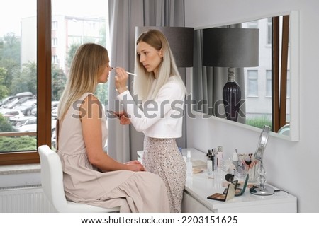 Make-up artist working with client in light beauty salon. Woman applying decorative cosmetics in front of mirror for blond hair model. Beautician working at makeup studio