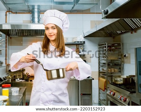 Woman cook. Girl works in restaurant kitchen. Young woman restaurant chef. Woman in chefs hat is holding price tag. Cooking in cafe kitchen. Chef career. Girl cook looking at camera. HoReCa industry