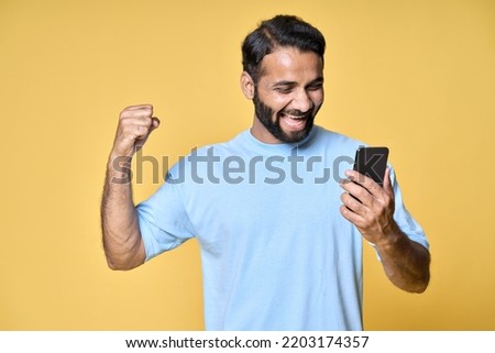 Excited overjoyed happy indian man raising fist screaming using smartphone winning mobile game, betting money prize holding cell phone celebrating victory isolated on yellow background.