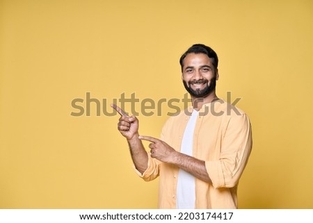 Happy indian man looking at camera pointing aside with fingers hand gesture indicating advertising new product commercial promotion, presenting sale offer standing isolated on yellow background. Royalty-Free Stock Photo #2203174417