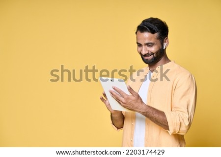 Happy indian man using digital tablet standing isolated on yellow background. Smiling ethnic guy holding pad wearing earbud watching virtual class webinar, having remote video call.