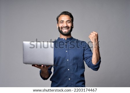 Happy excited indian business man employee holding laptop computer standing isolated on gray background celebrating win, good online results, business success, job promotion or achievement concept.