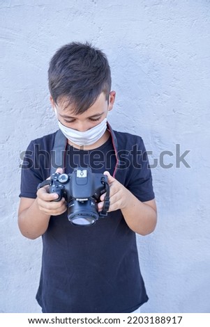 A cute little kid with green eyes wearing a mask is holding a camera and taking pictures . Happy boy dreams of becoming photographer.