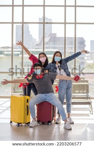 Happy Asian young friends with face mask raise hands to take photo at airport departure terminal. Travel with new normal to prevent covid-19 omicron pandemic.