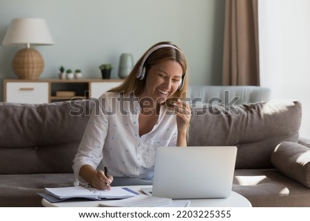 Happy joyful woman in wireless headphones talking on video call at laptop, laughing, having funny online conversation, writing notes, studying from home, watching webinar, class on Internet