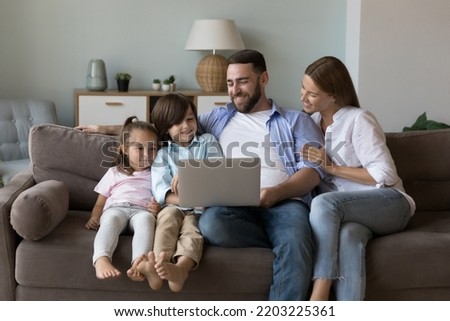 Happy family couple and cute cheerful kids using laptop on home couch, relaxing in living room together, watching movie with interactive app, Internet service. Domestic communication concept