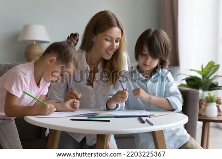 Happy mother helping cute little sibling kids to draw in colorful pencils in paper album, sitting at small table in living room, enjoying learning creative process, playtime, family leisure