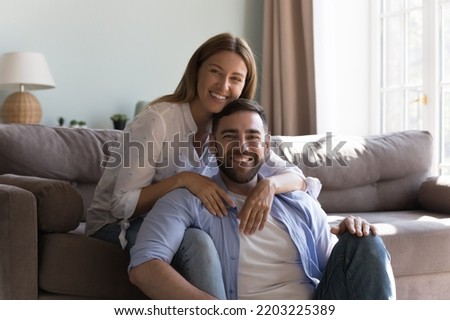 Happy attractive millennial couple posing in living room, resting on sofa and floor, hugging, looking at camera, smiling, dating at home. Marriage, relationship concept. Family portrait