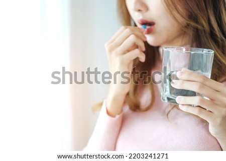 Sick Asian woman eating pills with a glass of water in hand near window in her house. Close up to a glass of water.