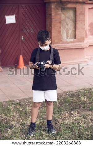 A cute little kid with green eyes wearing a mask is holding a camera and taking pictures . Happy boy dreams of becoming photographer.