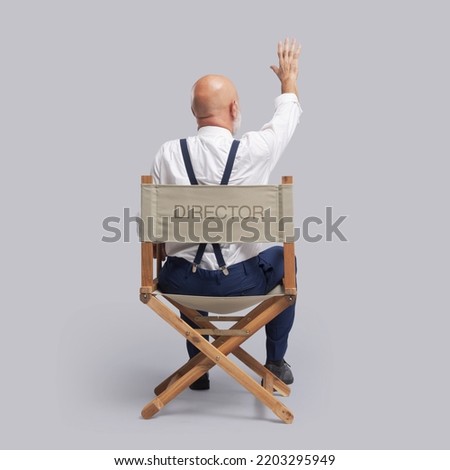 Professional filmmaker sitting on the director's chair and raising a hand, he is guiding the actors and the crew, back view Royalty-Free Stock Photo #2203295949