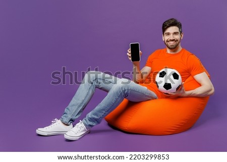 Full body young man wear orange t-shirt cheer up support football sport team hold soccer ball mobile cell phone blank screen workspace tv area sit in bag chair isolated on plain dark purple background