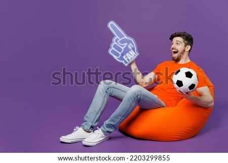 Full body young fan man wear orange t-shirt cheer up support football sport team hold soccer ball 1 fan foam glove finger up watch tv live stream sit in bag chair isolated on plain purple background