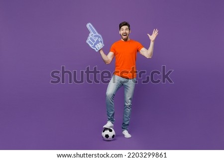 Full body young excited fun fan man he wear orange t-shirt cheer up support football sport team hold soccer ball fan foam glove finger up watch tv live stream isolated on plain dark purple background Royalty-Free Stock Photo #2203299861
