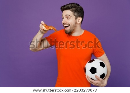 Young excited amazed fan man wears orange t-shirt cheer up support football sport team hold in hand soccer ball eat slice of italian pizza watch tv live stream 2024 isolated on plain purple background