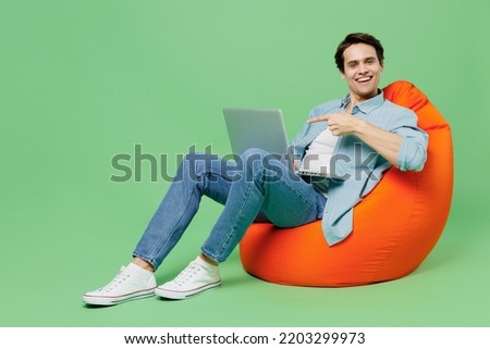 Full size fun young brunet man 20s wears blue shirt sit in bag chair hold use work on laptop pc computer pointing forefinger on screen looking camera isolated on plain green background studio portrait