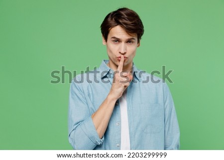 Secret calm sullen frowning gloomy young brunet man 20s years old wears blue shirt say hush be quiet with finger on lips shhh gesture looking camera isolated on plain green background studio portrait Royalty-Free Stock Photo #2203299999