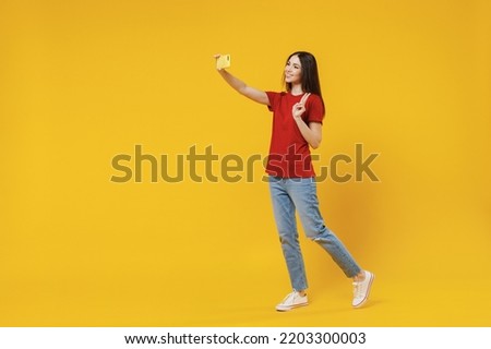 Full size body length young brunette woman 20s wears basic red t-shirt do selfie shot on mobile cell phone post photo on social network show victory sign isolated on yellow background studio portrait.