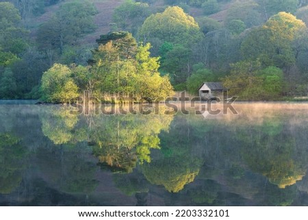 Beautiful Spring colours on trees at Rydal Water in the Lake District, UK. Perfect reflections in misty water with small boathouse. UK landscape photography. Royalty-Free Stock Photo #2203332101