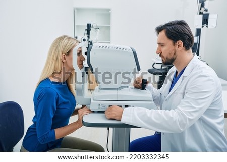 Experienced ophthalmologist man using automated refractor for vision correction female patient in ophthalmic office Royalty-Free Stock Photo #2203332345