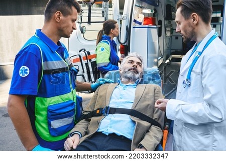 Paramedic and doctor examining of senior male patient lying in ambulance gurney brought to hospital by EMT workers. Emergency medical services and caring about person Royalty-Free Stock Photo #2203332347