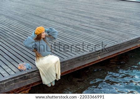 Outdoors fashion portrait of a beautiful middle aged woman walking on the beach. Marine background. Dressed in a stylish warm blue sweater, yellow skirt and beret.