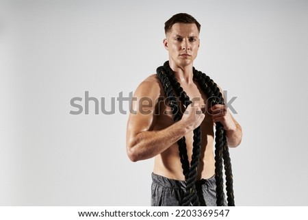 Confident young man with perfect torso carrying rope on shoulders against white background