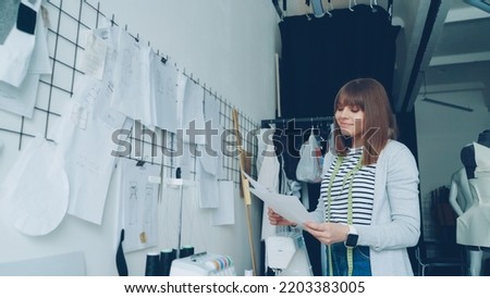 Young creative clothes designer is looking at sketches then hanging them on wall above studio table beside other drawings. Loft style studio and fashionable garments are visible.
