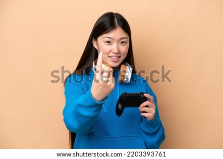 Young Chinese man playing with a video game controller isolated on beige background doing coming gesture