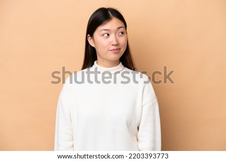 Young Chinese woman isolated on beige background making doubts gesture looking side