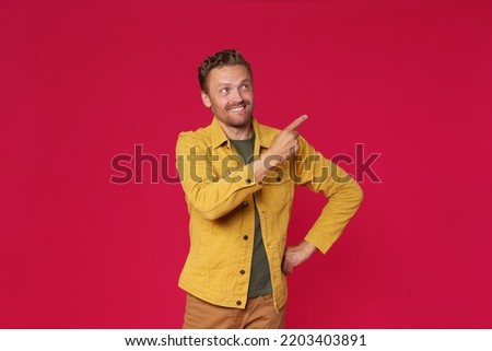 Handsome young man in jeans jacket pointing looking sideways up mock up advertisement isolated on red background. Charming smiling freelancer introduce your business offer.  Royalty-Free Stock Photo #2203403891