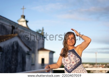 A woman on the porch of her house looking at the camera against the sky and church in the background. City of Valença, Bahia, Brazil.