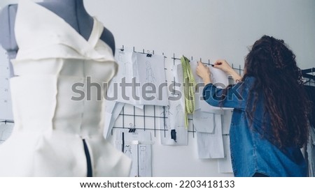 Young female clothing designer is looking at sketches and hanging drawings on wall in light workshop. Large collection of drafts above tailoring desk, clothed dummy in foreground.