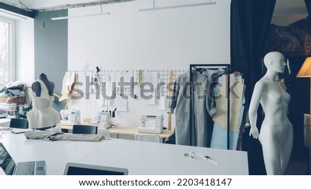 Light clothing design studio with large tailor's desk, mannequins, numerous sketches pinned on wall, sewing machines, tailoring items and half-finished garments on rails.