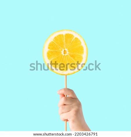 Minimal creative food concept made with child’s hand holding stick with lemon slice, against light, vibrant blue background. The idea of healthy sweets or citrus lollipop. Natural source of vitamin C.