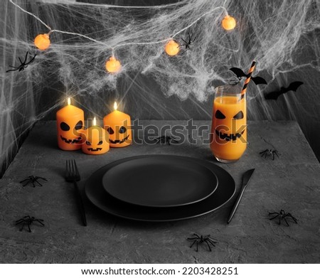 Halloween table setting with black cutlery. Festive serving with burning candles and garland, pumpkin juice, spiders on a dark gray background. Empty plate.