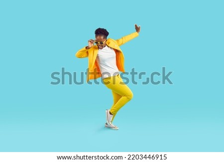 Cheerful funny woman in good mood dancing cheerfully isolated on light blue background. Young stylish African American woman in yellow casual suit, t-shirt, sneakers and sunglasses. Full length. Royalty-Free Stock Photo #2203446915