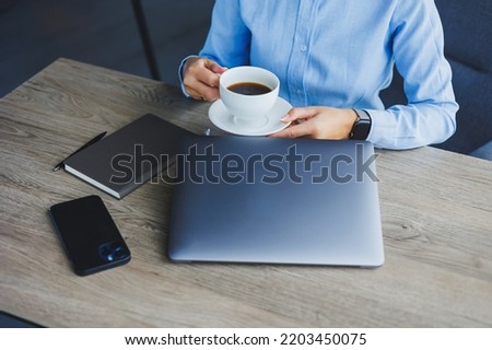 Portrait of cheerful woman in classic glasses smiling in free time in cafe with coffee, positive european woman in blue shirt, desk with laptop, remote work