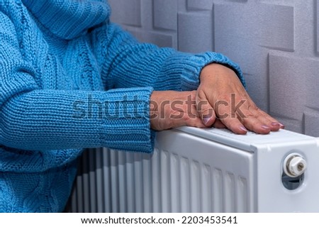 An elderly woman in a blue sweater warming her hands on a heating radiator. Concept: low room temperature, rising gas and heating prices, getting colder in the apartment. Royalty-Free Stock Photo #2203453541