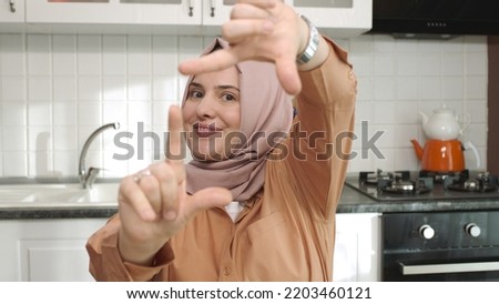Woman in hijab sitting in kitchen making frame gesture. She is looking at the camera and framing the camera with her hands.
