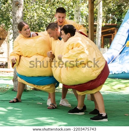 Young and adult men and women in large sumo suits pushing each other, having fun in adventure park.