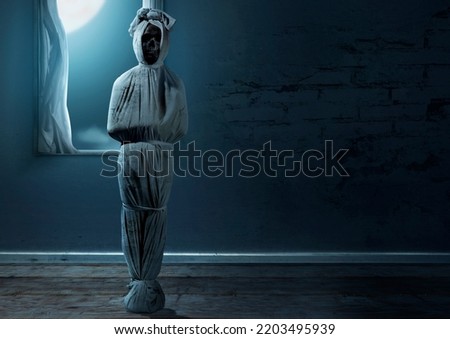 Pocong is covered with a white linen shroud standing on an abandoned house. Halloween concept Royalty-Free Stock Photo #2203495939