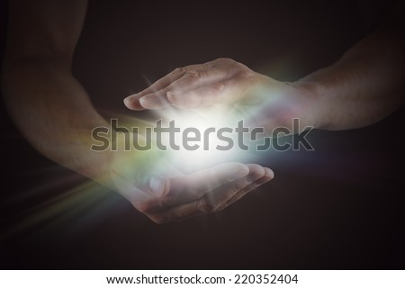 The Spark of Life - Male hands emerging from darkness, cupped, with rainbow colored bright white star burst of light between Royalty-Free Stock Photo #220352404