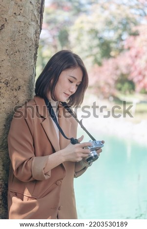 Vertical Asian Women with vintage film camera take a photo. Smiling female photographer look at photo from professional camera outdoor. Vertical photo of young woman shoot photo in green nature park