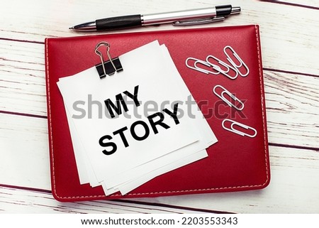 On a light wooden background, a burgundy pen and notebook. On the notebook has white paper clips and white paper with the text MY STORY. Business concept