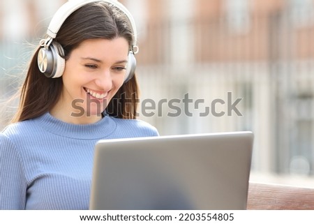 Happy woman using laptop with headphones sitting on a bench in the street