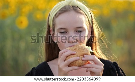 Teen girl eats hamburger enjoying meal against blurry sunflower field on nature