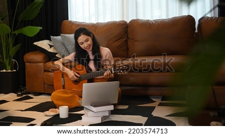 Young woman learning to play guitar, using laptop computer on carpet in living room Royalty-Free Stock Photo #2203574713