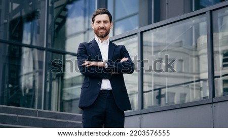 Successful confident caucasian bearded businessman in suit standing outdoors on background office building smiling proud male professional worker leader looking at camera posing with crossed arms